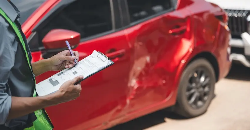 An insurance adjuster in a green safety vest writes on a clipboard while standing beside a red sedan with significant door damage depicting the on-site vehicle assessment process that kicks off car insurance claims after a collision in North Carolina.