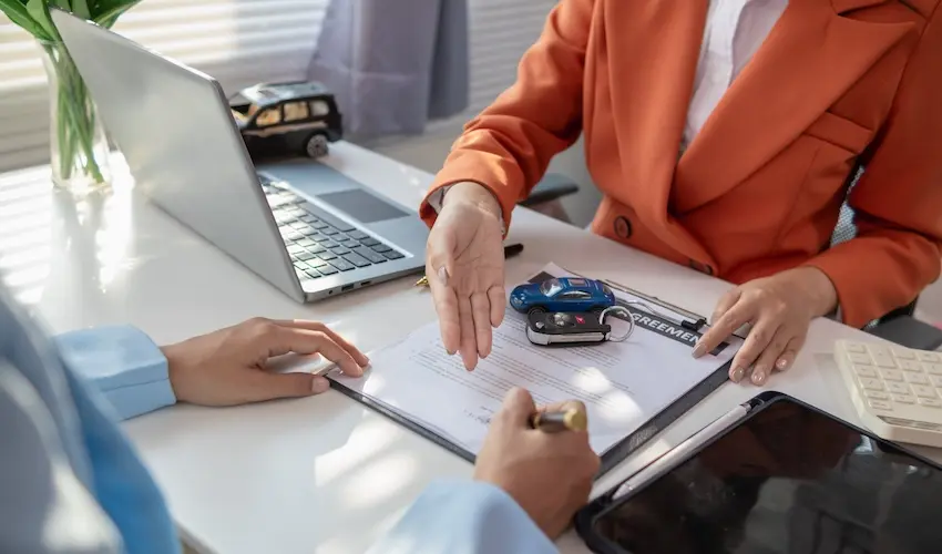 A woman in an orange blazer slides a printed agreement document across a desk toward a client while car keys and a laptop sit nearby portraying the formal paperwork review and settlement process involved in resolving car insurance claims and legal compensation with help from Mogy Law.