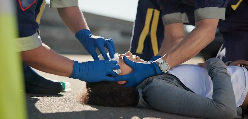 Two paramedics wearing blue latex gloves kneel over a young woman lying unconscious on the pavement carefully stabilizing her head and neck while administering emergency treatment depicting first responders managing some of the most critical car accident injury types including spinal and head trauma at the scene of a collision in North Carolina.