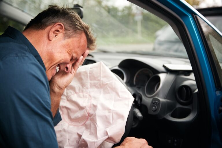 An older man with gray hair slumped forward in the driver's seat of a blue car with a deployed airbag pressing his hand to his face in pain and distress through the open door illustrating the facial- chest- and head car accident injury types that drivers commonly sustain when an airbag deploys during impact in North Carolina.