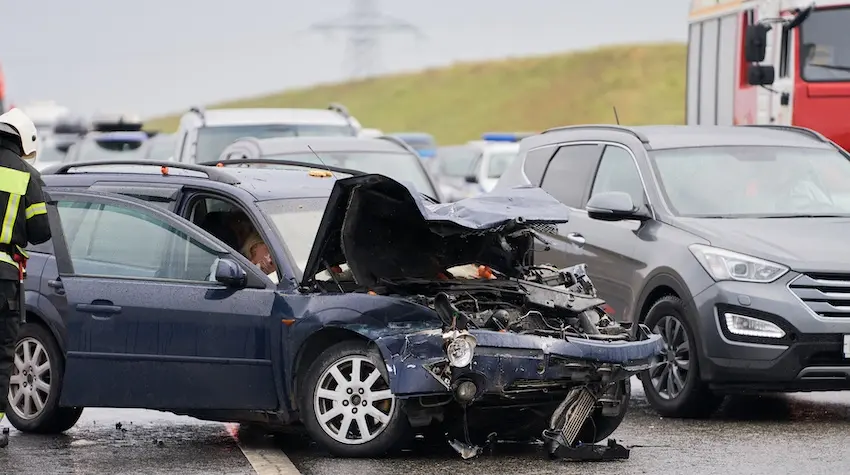 A blue sedan with its hood completely crushed and engine fully exposed sits in the middle of a rain-soaked highway surrounded by other vehicles while a firefighter in a white helmet and yellow vest approaches with a red emergency truck and police cars visible in the background representing the multi-vehicle pileup scenario that produces the most severe and varied car accident injury types in North Carolina and could use representation from Mogy Law.