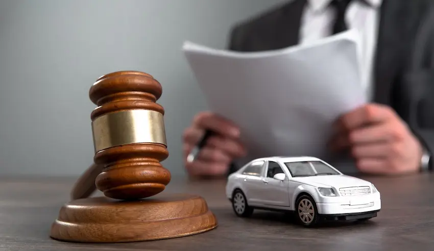 A wooden judge's gavel with a gold band and a white toy sedan sits on a desk in the foreground while a lawyer in a dark suit from Mogy Law reviews a stack of legal documents in the background representing the legal process of seeking compensation for the full range of car accident injury types sustained in a collision.
